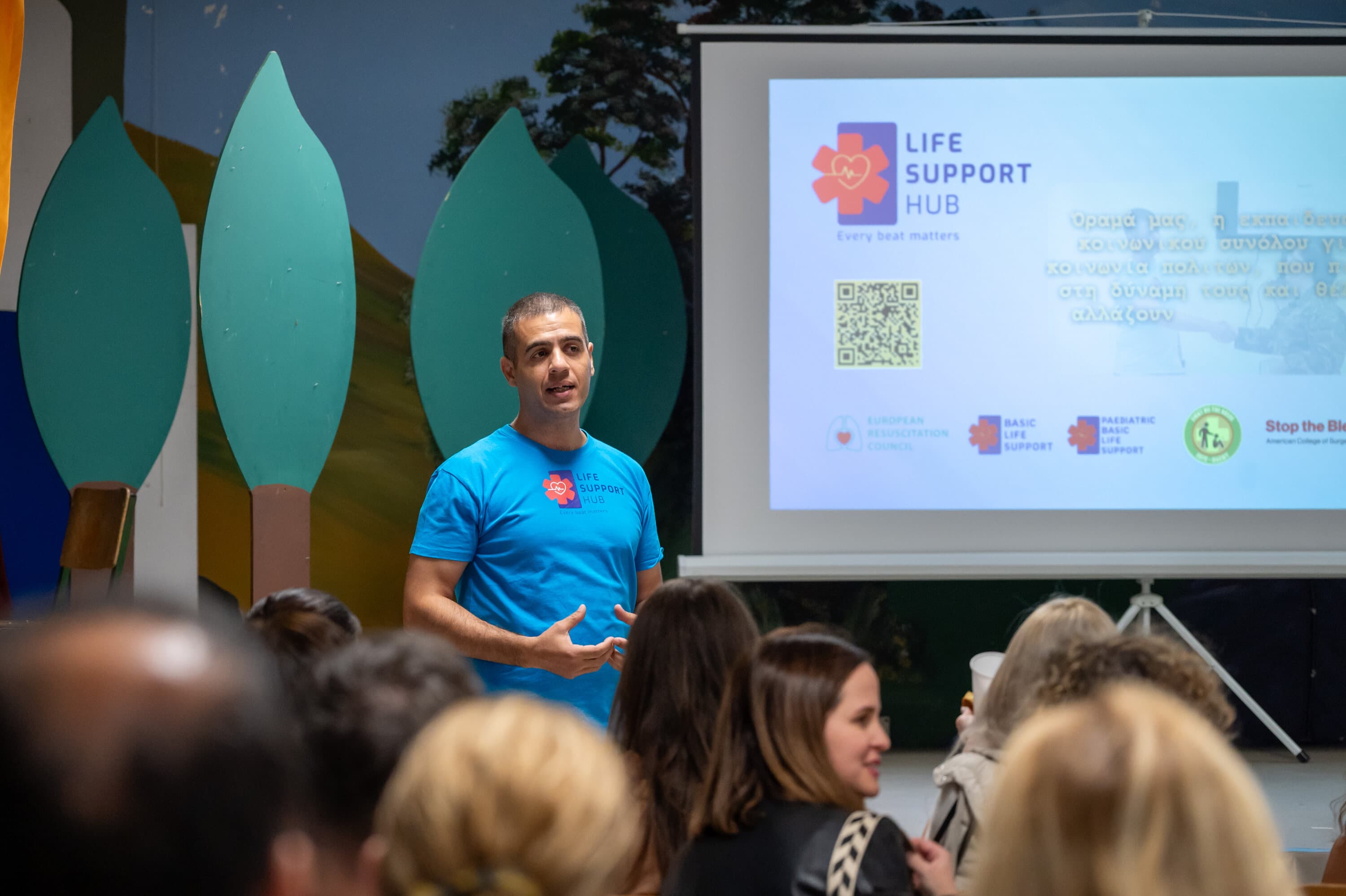 Εκπαιδευτής της Life Support Hub μιλά σε ακροατήριο· στην οθόνη το λογότυπο και οι πιστοποιήσεις του οργανισμού